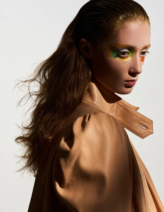 Close-up portrait of model Lydia Reid with colorful eye makeup and wet-look hair wearing a camel trench coat against white seamless background