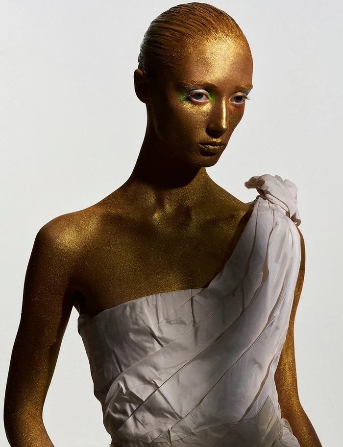 Model with metallic gold body paint and white dress against a plain white background for Vogue Greece