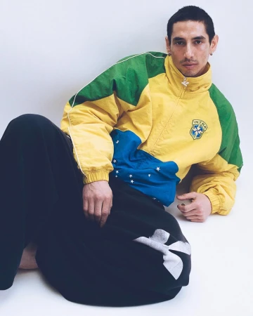 Man in a yellow, green, and blue Intra zip windbreaker and black pants reclining against a white studio backdrop.