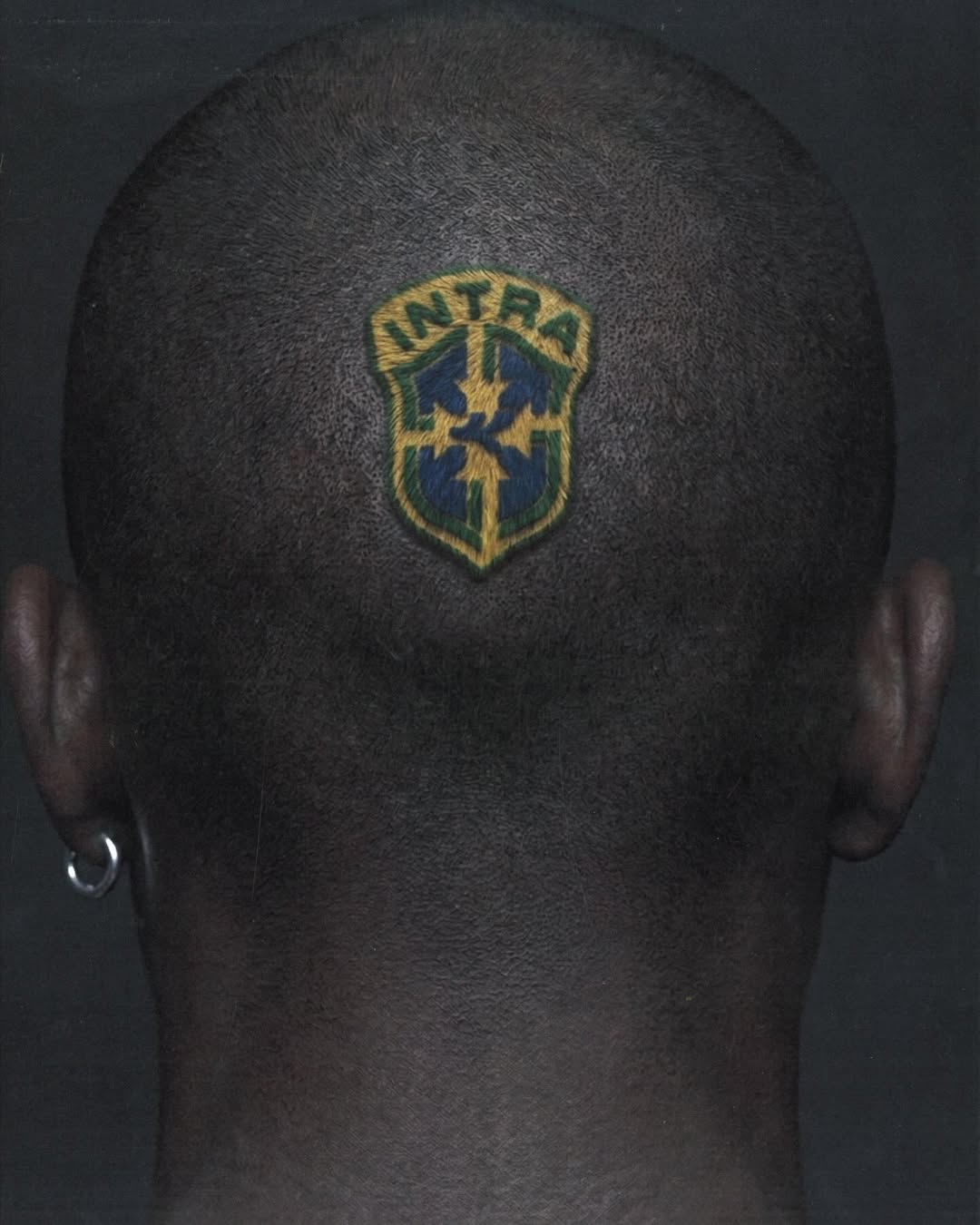 Back of a person's shaved head featuring an Intra crest dyed into the buzzcut, wearing a silver hoop earring against a dark background.