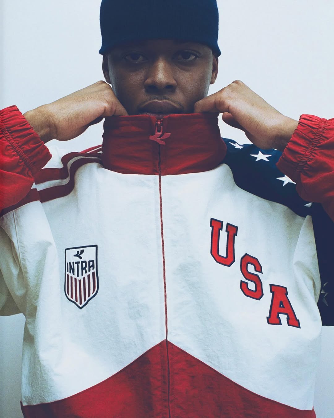 Portrait of a man wearing a red, white, and blue Intra USA track jacket and dark beanie against a plain backdrop.