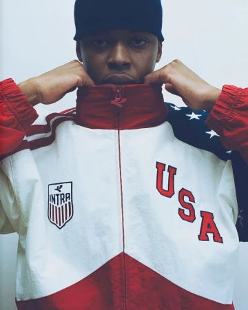Portrait of a man wearing a red, white, and blue Intra USA track jacket and dark beanie against a plain backdrop.