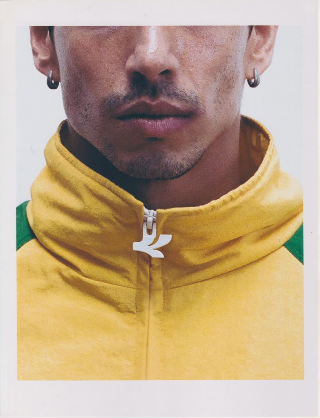 Close-up portrait of a man wearing small hoop earrings and a yellow zipped track jacket with green accents against a white background.