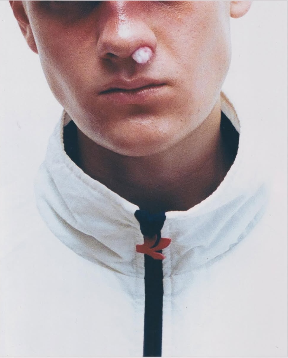 Close-up of a young man wearing a white zip-up jacket with cotton in his nostril against a plain white background.