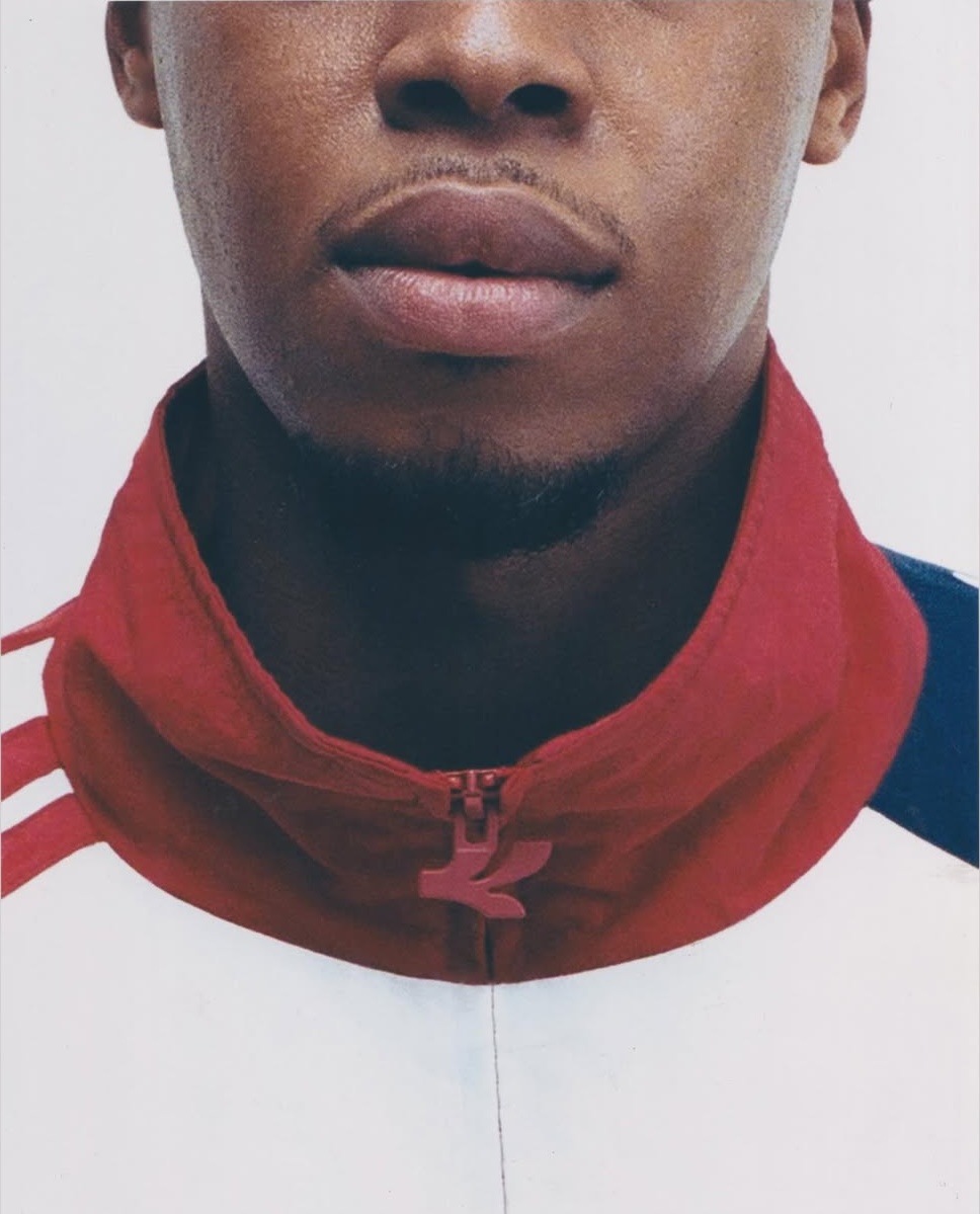Close-up crop of a Black man wearing a white and red zip-up athletic jacket against a white background.