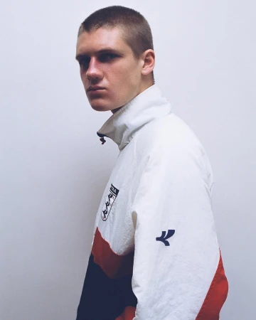 Young man with a buzz cut wearing a white, red, and navy zip-up sports windbreaker against a white background.