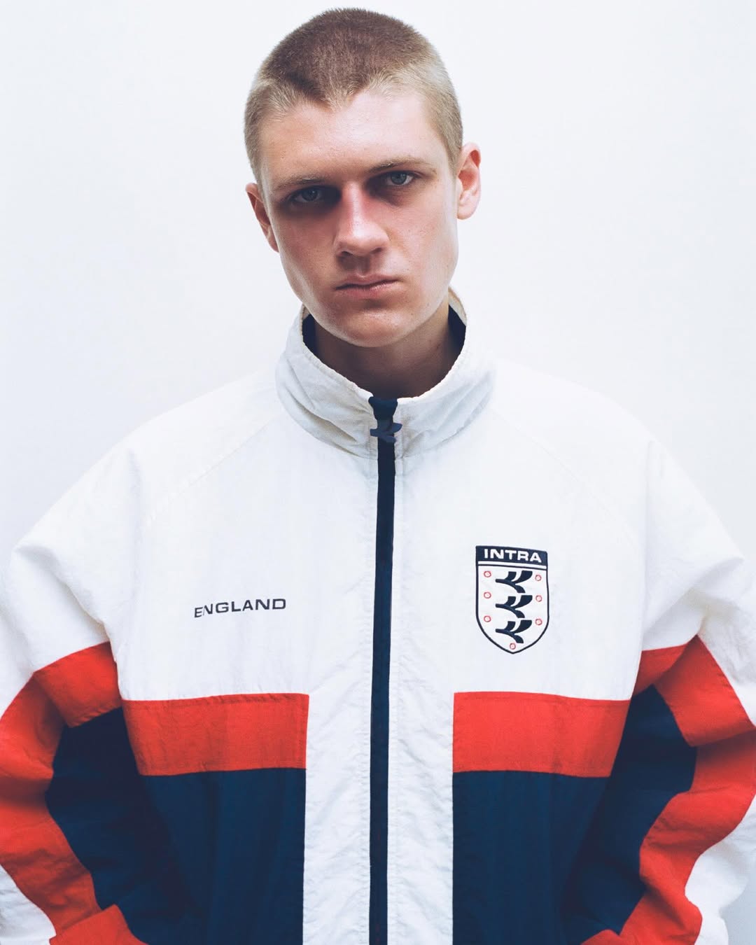 Young man with buzzcut hair wearing a white, red, and navy blue Intra England track jacket against a white studio background.
