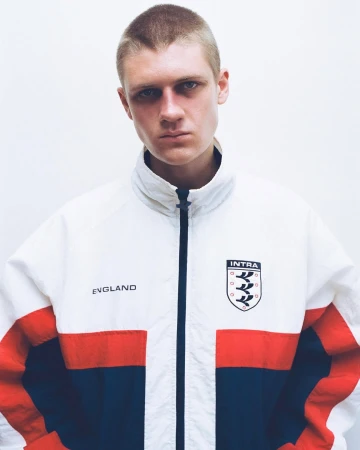 Young man with buzzcut hair wearing a white, red, and navy blue Intra England track jacket against a white studio background.