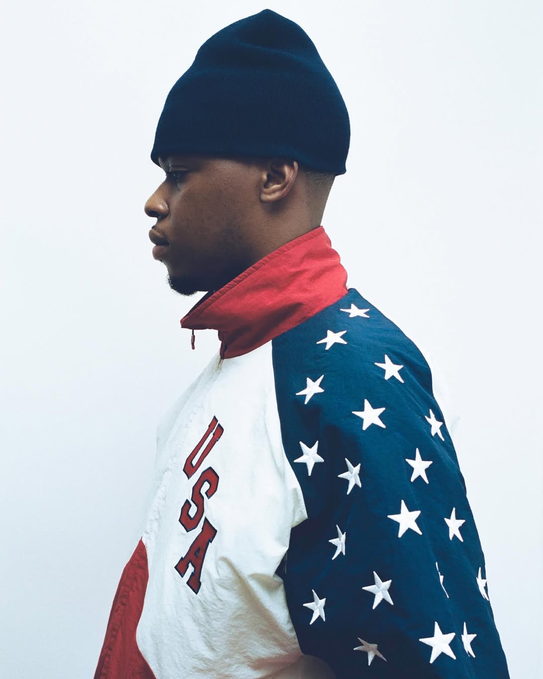 Young Black man in profile wearing a dark beanie and a vintage USA star-patterned windbreaker jacket against a white studio background.