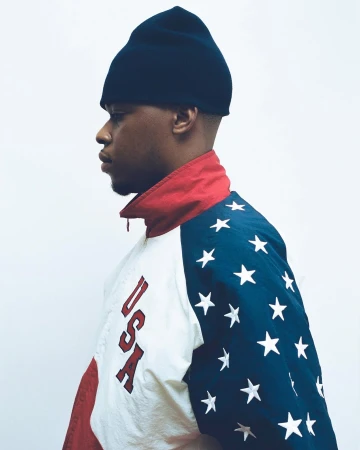 Young Black man in profile wearing a dark beanie and a vintage USA star-patterned windbreaker jacket against a white studio background.