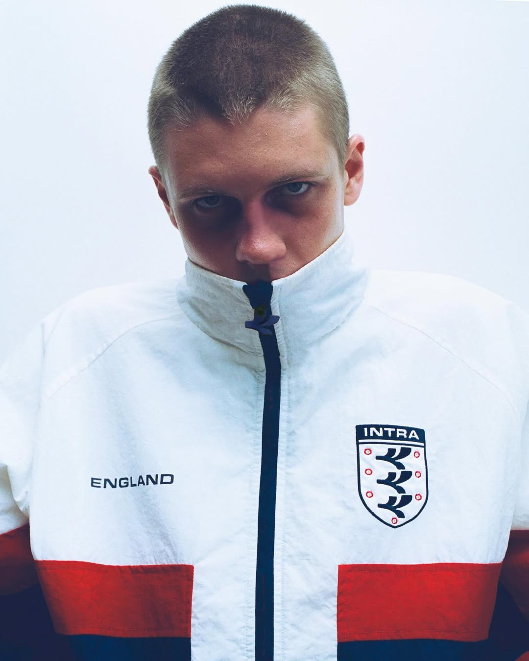 Buzzcut young man wearing a white, red, and navy Intra England track jacket looking intensely at the camera on a white backdrop.