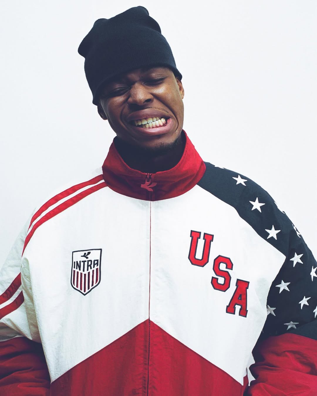 Black man wearing a black beanie and retro red, white, and navy USA Intra track jacket against a white studio background.