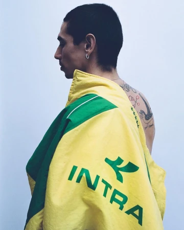 Man in profile wearing a yellow and green Intra track jacket over bare tattooed shoulders against a light background.