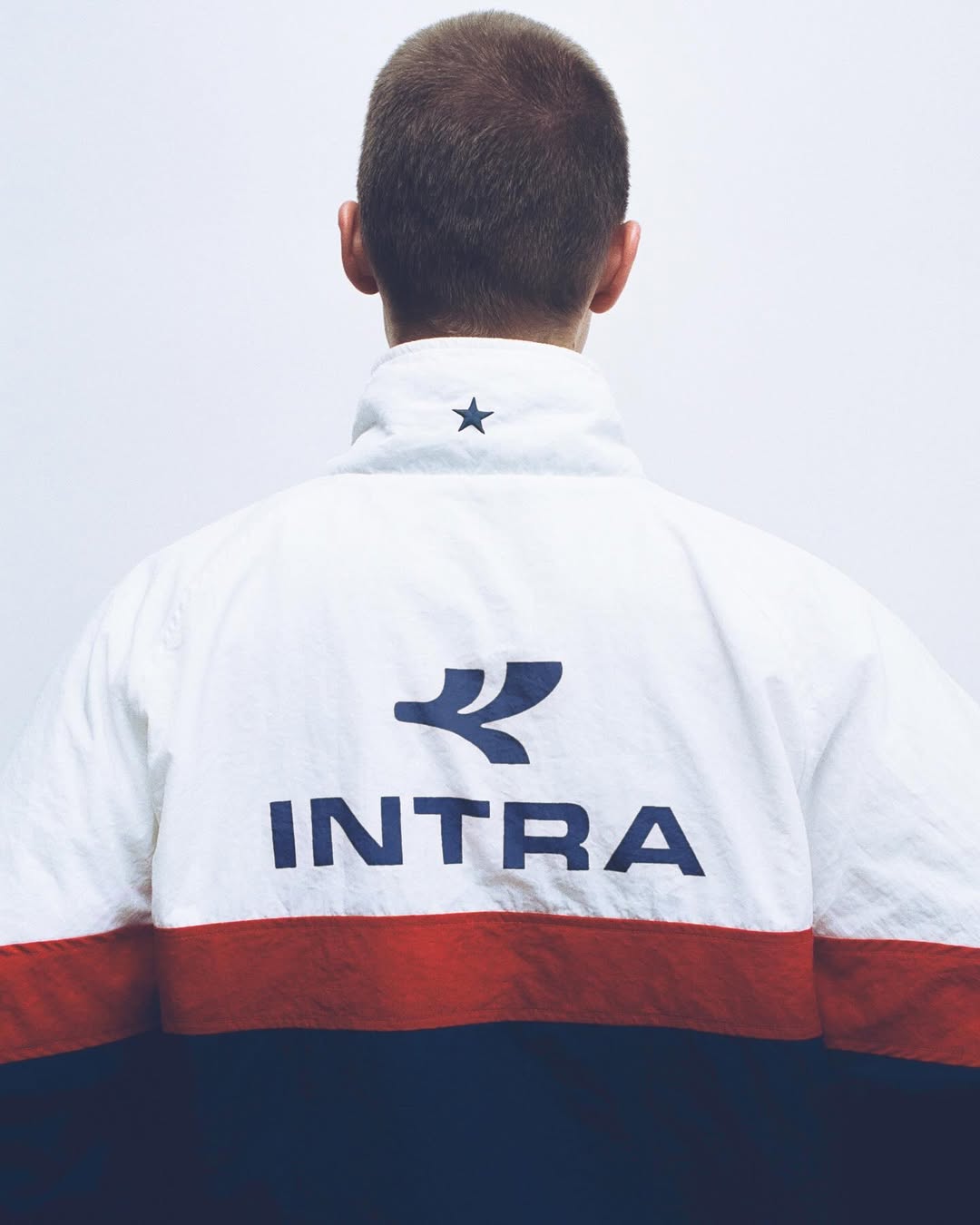 Back view of a man wearing a white, red, and navy Intra windbreaker jacket against a white studio background.
