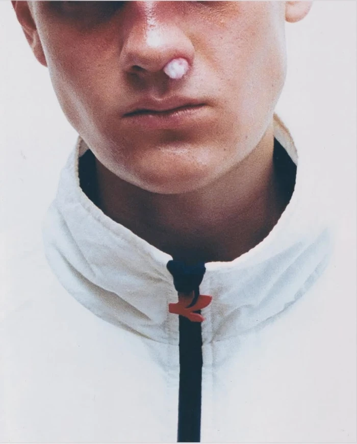 Close-up of a young man wearing a white zip-up jacket with cotton in his nostril against a plain white background.