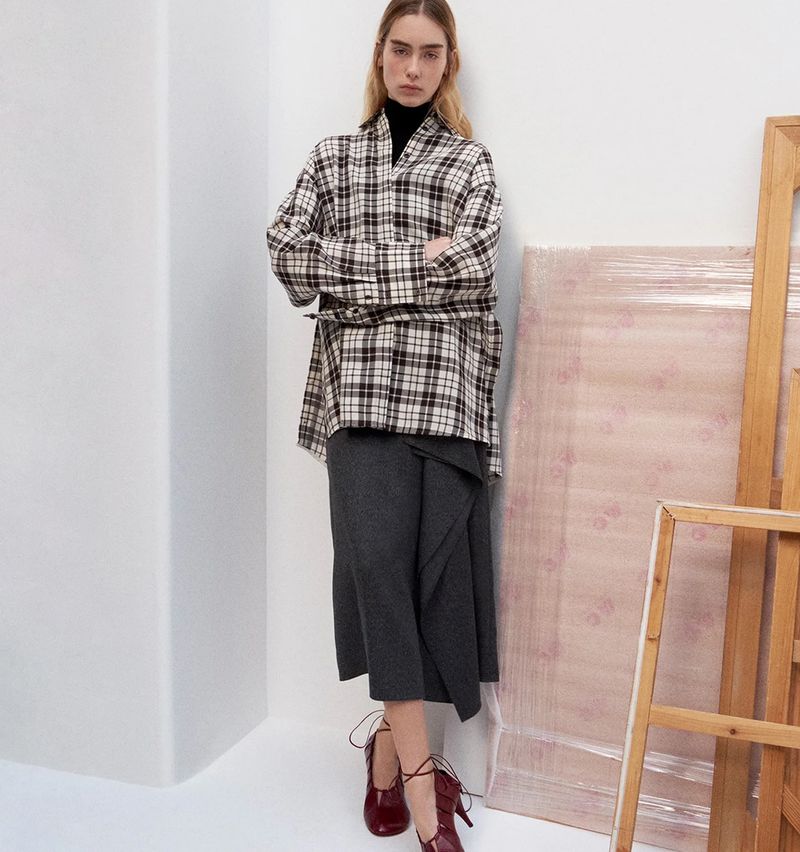 A woman poses in a plaid shirt, black turtleneck, and grey skirt against a white corner wall with painting materials in the background.