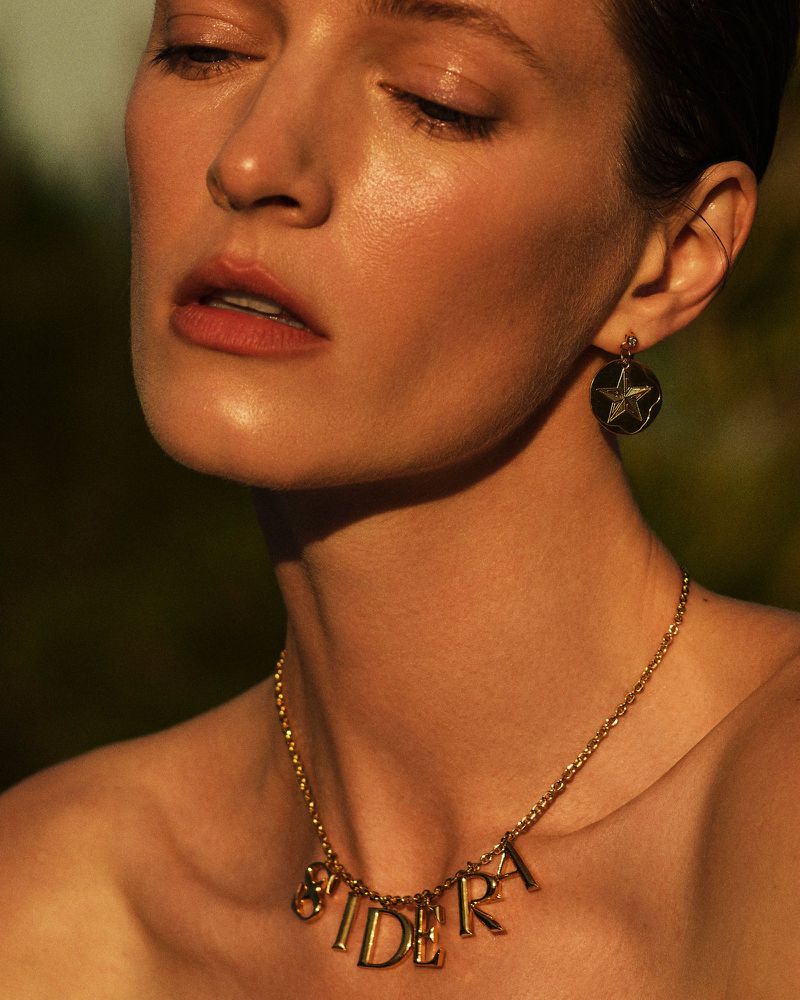 Close-up of a woman wearing a gold SIDERA letter necklace and a round star earring in warm golden sunlight.