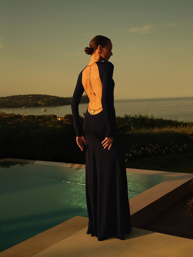 Model in a dark backless gown with delicate gold back jewelry standing beside a swimming pool overlooking a coastal bay at sunset.