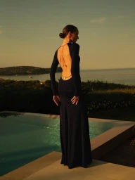 Model in a dark backless gown with delicate gold back jewelry standing beside a swimming pool overlooking a coastal bay at sunset.