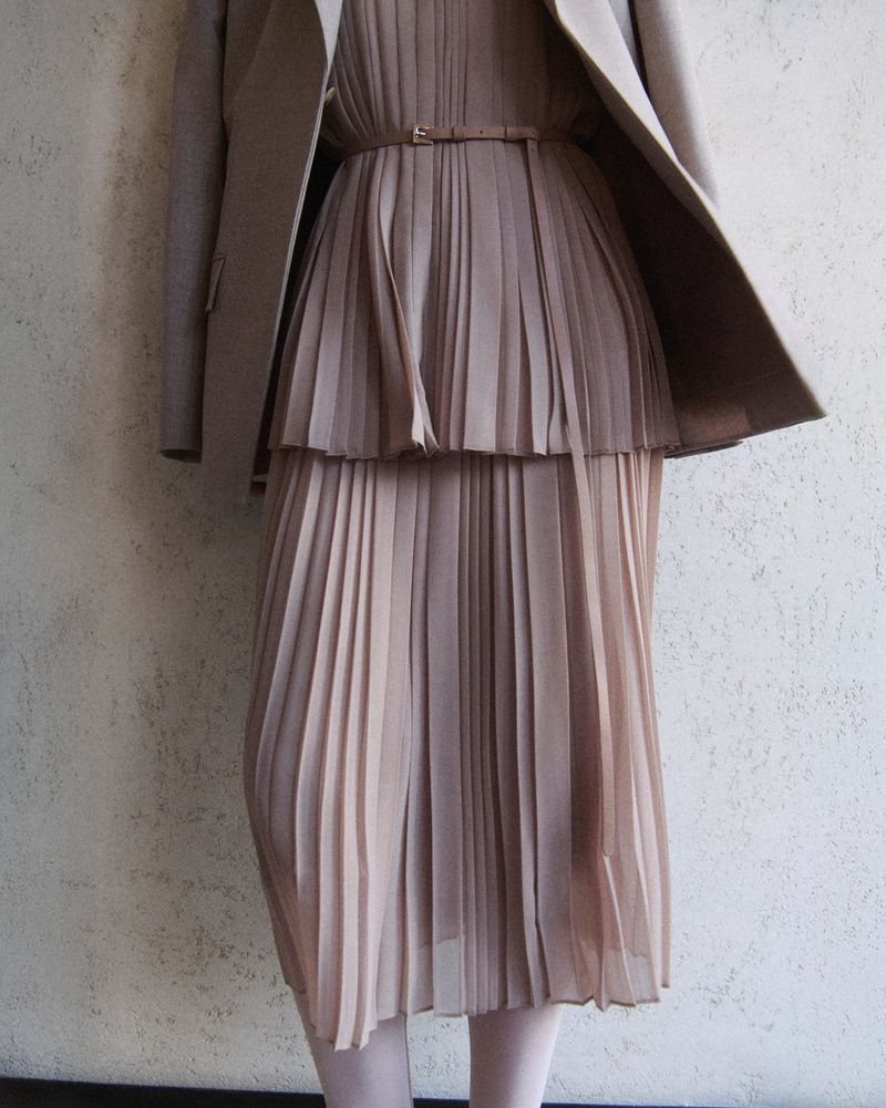 Mid-section shot of a fashionably dressed person in a beige pleated dress, cinched with a belt, complemented by a matching blazer.