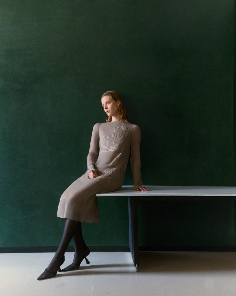 A fashion shot of a woman in a tan dress with dark tights sitting on a table in front of a dark green wall with studio lighting.