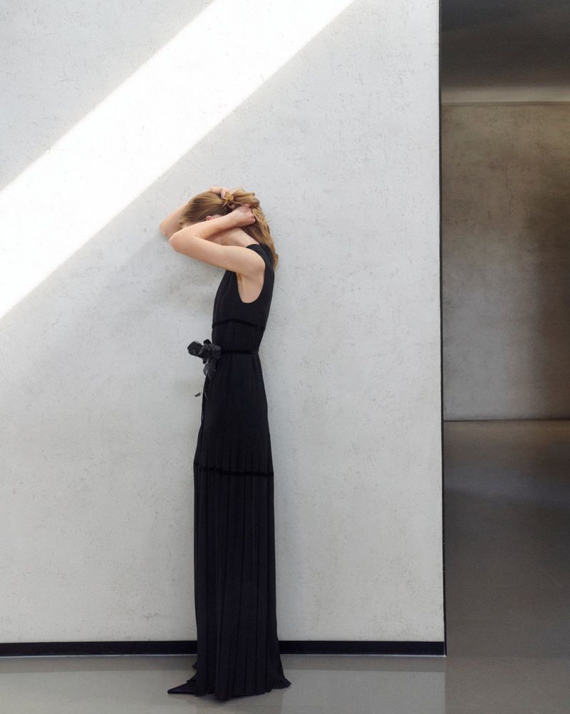 Woman in a black dress stands against a textured white wall with strong light from above, creating shadows in an architectural interior.