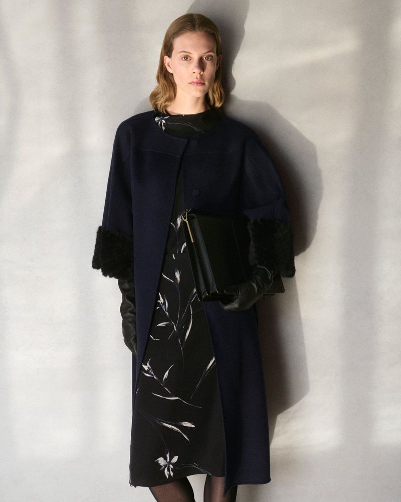 A woman in a navy coat with fur cuffs and a floral dress poses indoors with a black bag. Fashion-forward studio portrait.
