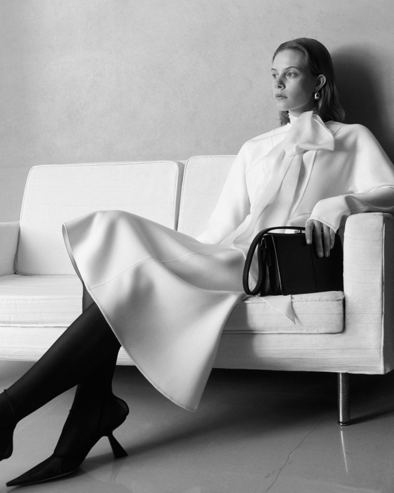 Model Anouk Smits in a white tie-neck dress, black tights, and heels seated on a sofa with a black handbag for Giada.