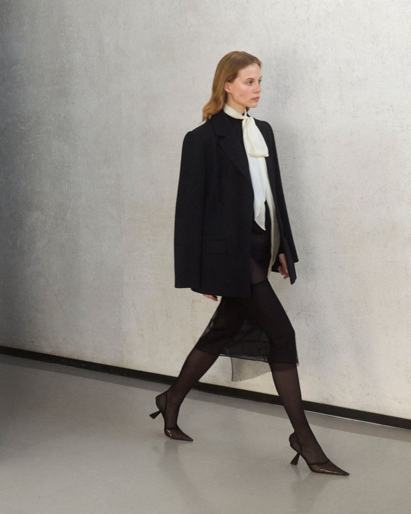 Woman in black blazer and sheer skirt walking on a runway with high heels. Fashion model presenting clothing during a show.
