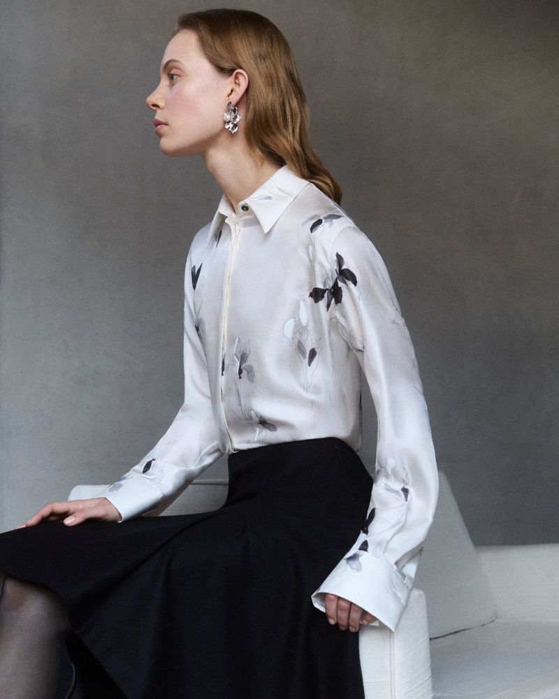 A woman with light brown hair poses in a button-down shirt with butterfly prints and a black skirt against a gray background.