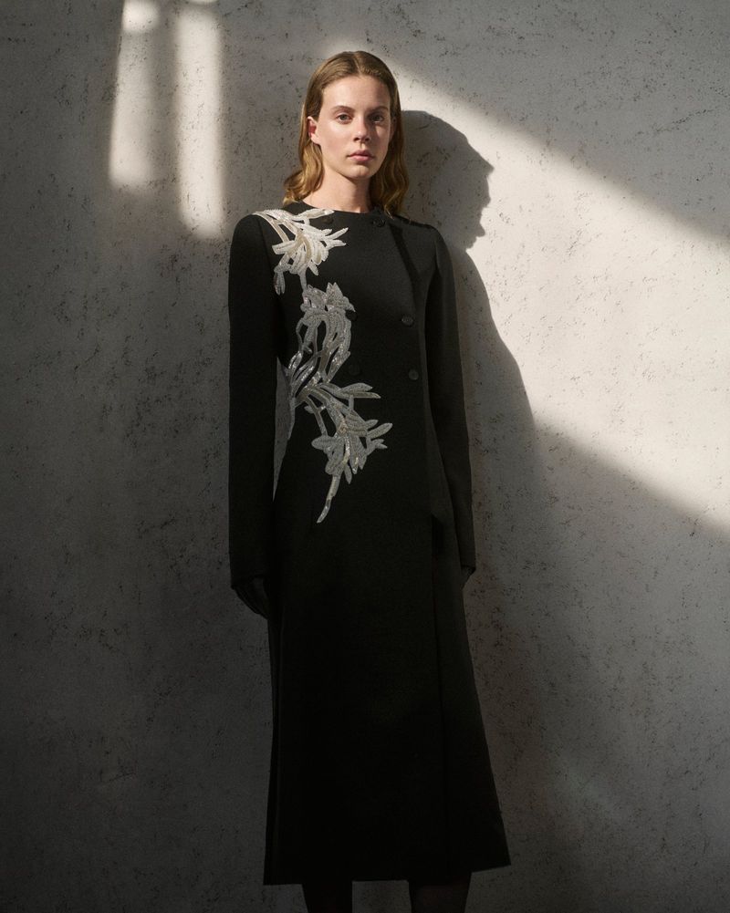 Woman in a black dress with silver floral embellishments posing against a textured wall with a beam of light creating shadows.