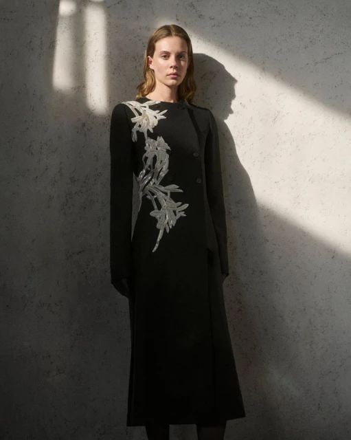 Woman in a black dress with silver floral embellishments posing against a textured wall with a beam of light creating shadows.