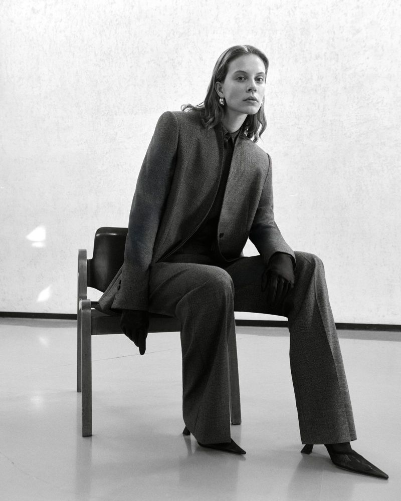 Adult woman in a grey suit sitting on a chair in a minimalist studio setting, captured in a medium shot emphasizing fashion and poise.