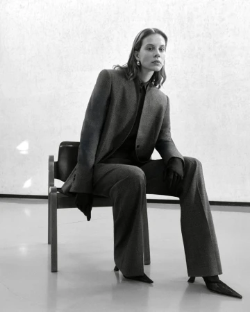 Adult woman in a grey suit sitting on a chair in a minimalist studio setting, captured in a medium shot emphasizing fashion and poise.