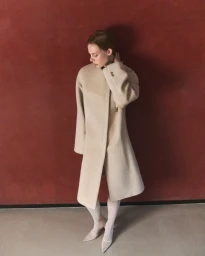 Model in cream coat wearing white tights standing against a textured red wall