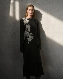 Model Anouk Smits wears an embroidered black long-sleeve dress against a textured wall with sunlight and shadows for Giada.