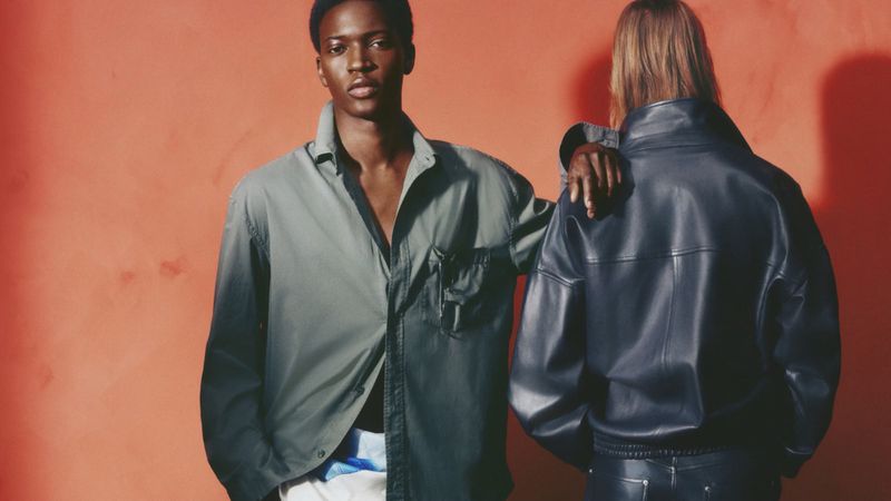 A Black male model in an olive button-down shirt rests his hand on the shoulder of a model in a dark leather jacket against a red backdrop.
