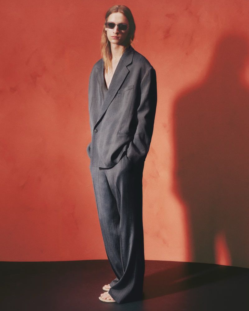 Model in grey oversized suit and sunglasses standing against a textured red backdrop for H&M Atelier