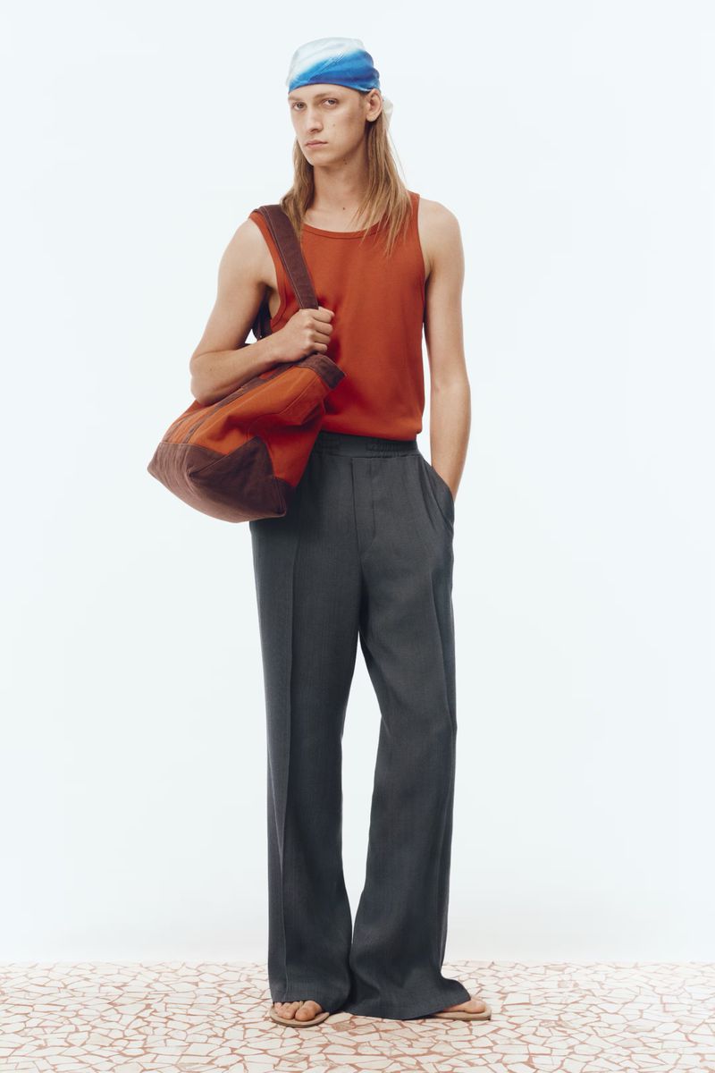 Model wearing an orange tank top, wide-leg charcoal trousers, a blue headscarf, and carrying a canvas tote bag against a white backdrop.