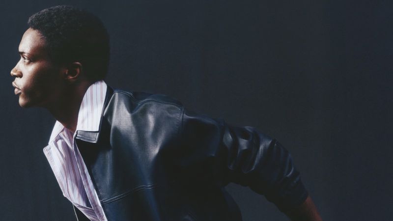 Black male model in profile wearing a black leather jacket and striped collared shirt against a dark background.