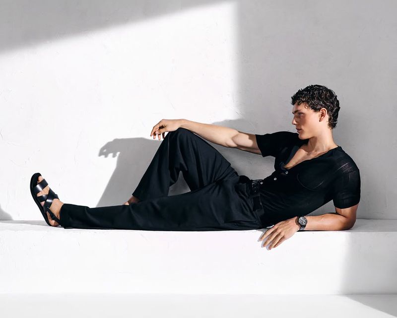 Attractive man lying on steps in black casual outfit and sandals, lit by soft natural light against a white background in a studio.