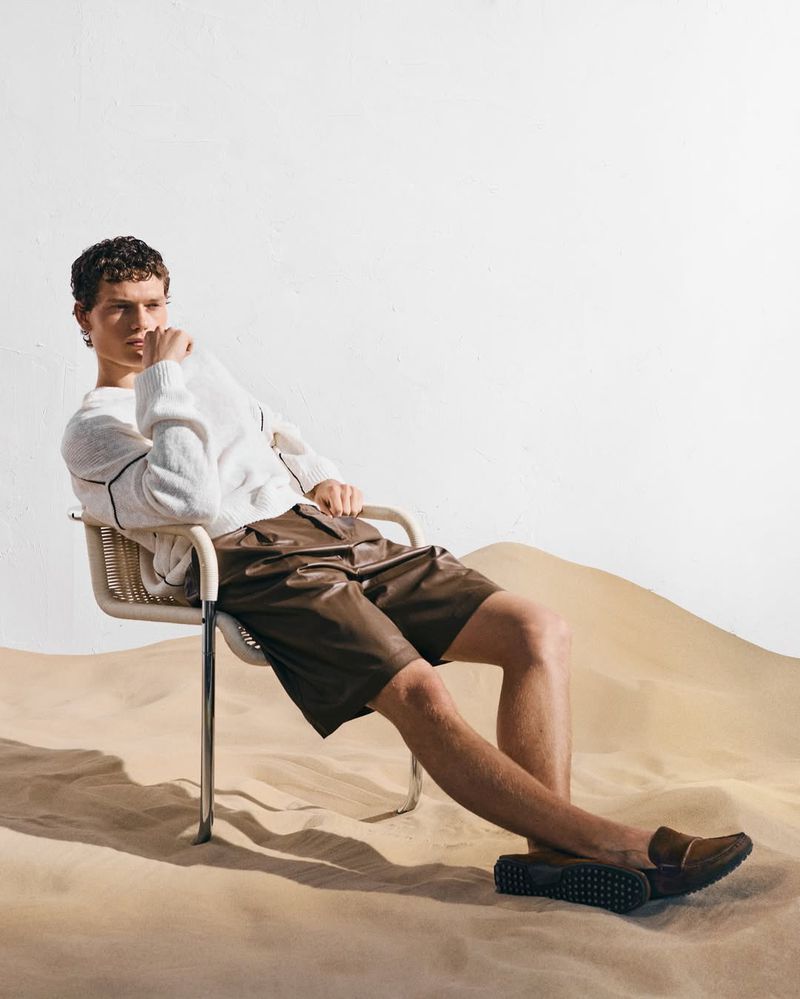 Model Caesar van den Idsert sitting in a modern chair on sand wearing a white knit sweater and brown leather shorts.