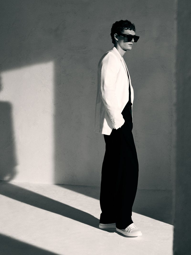 Monochrome fashion shot: Stylish adult male in a light jacket and dark trousers, standing indoors with strong shadows and wearing sunglasses.