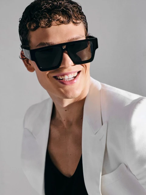 Smiling man with curly hair wears black sunglasses and a white blazer in a studio portrait against a neutral background.