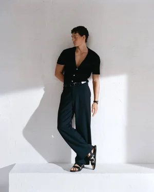 Male model wearing black shirt and dark trousers leaning against a white textured wall