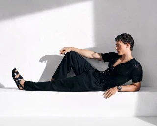 Model Caesar van den Idsert in black polo shirt and trousers lounging against white studio wall for Kazar SS'25 campaign.