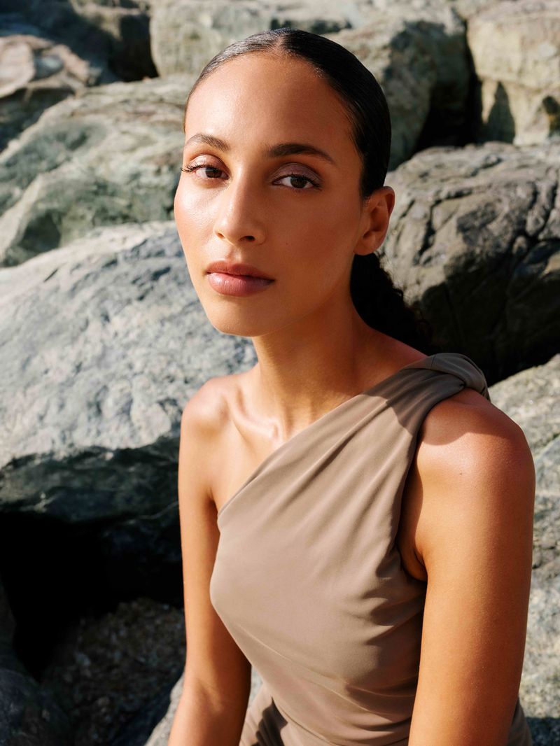 Portrait of a young woman with tanned skin wearing a brown, one-shoulder dress, posing against a background of grey rocks in natural light.