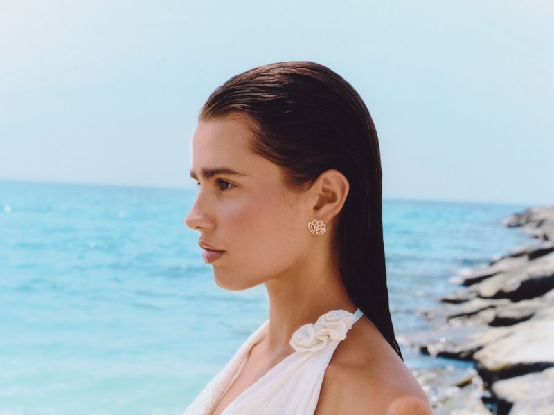 Profile shot of an adult female with dark hair wearing lotus earrings and a cream top against an ocean and blue sky backdrop.