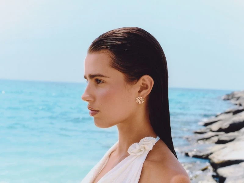 Profile shot of an adult female with dark hair wearing lotus earrings and a cream top against an ocean and blue sky backdrop.