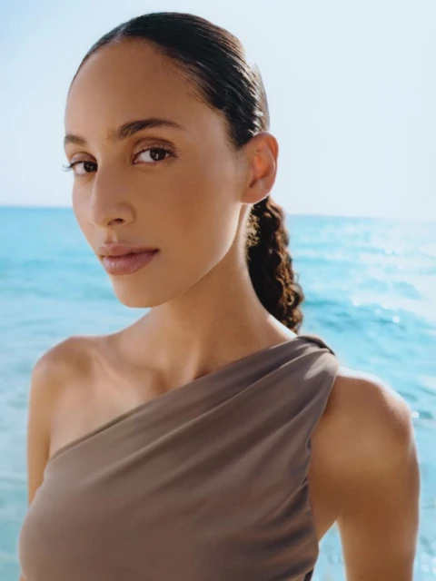 Portrait of a woman in a draped top against a bright blue ocean backdrop, capturing a serene and natural beauty moment in soft light.